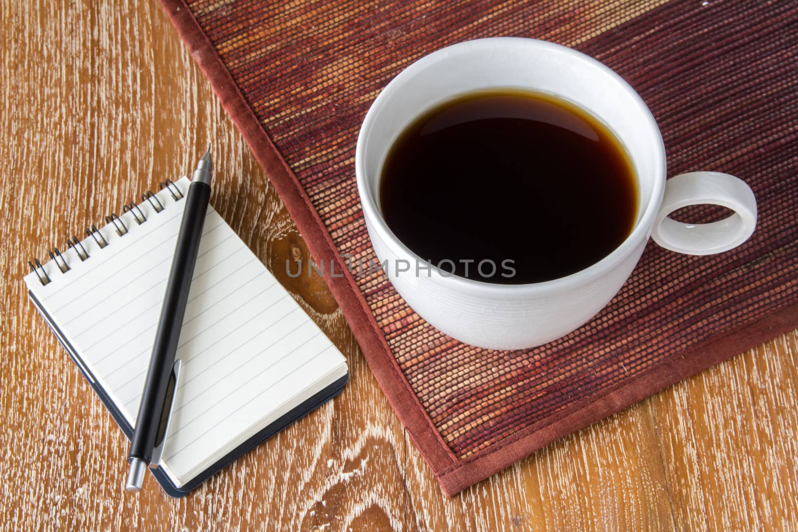 White cup of coffee on wooden table with mat and notebook