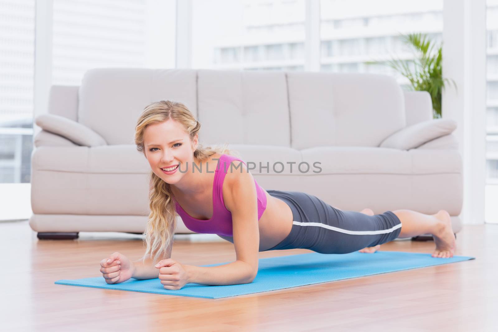 Fit blonde in plank position on exercise mat smiling at camera at home in the living room