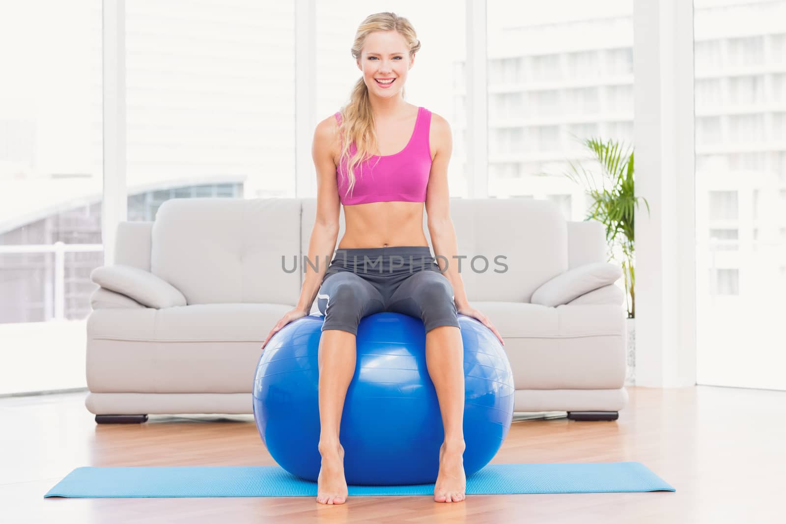 Fit blonde sitting on exercise ball smiling at camera at home in the living room
