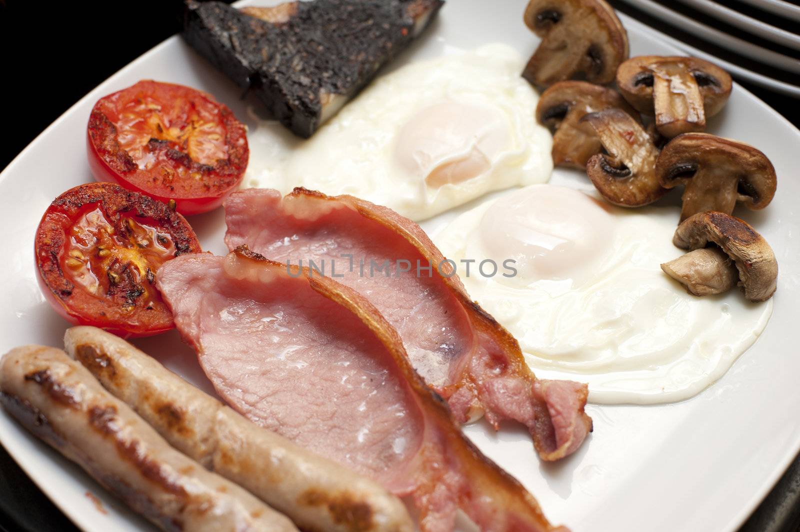 Full cooked English breakfast with a serving of fried eggs, bacon, tomato, sausages, hash brown and mushrooms, close up view on a plate