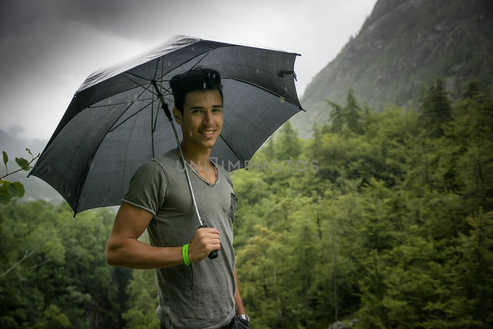 Smiling handsome young man holding an umbrella on a stormy day as he takes a walk along a trail in the mountains turning to smile at the camera