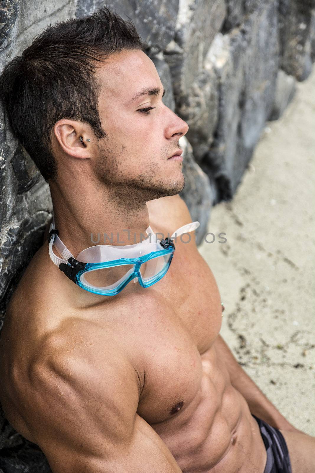 Handsome muscular fit young man relaxing at the beach sitting on the sand leaning against a wall with his swimming goggles around his neck, closeup portrait in profile