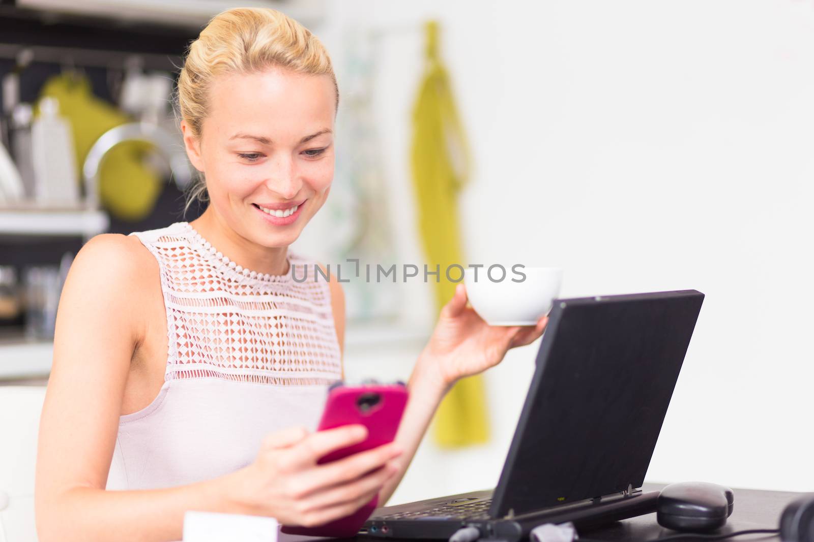 Business woman working from her home. Multitasking. Having morning coffee while texting on the smarth phone and working on leptop computer.