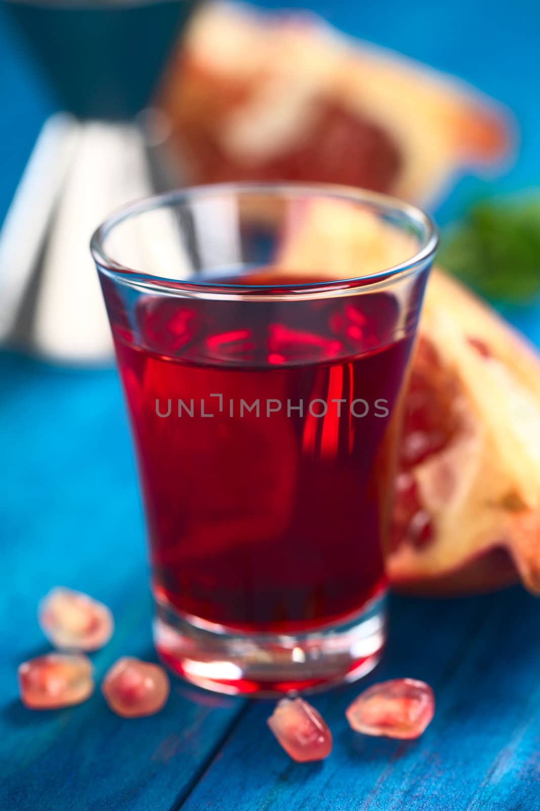 Grenadine syrup in shot glass with pomegranate seeds in the front and pomegranate in the back (Selective Focus, Focus on the front rim of the glass)