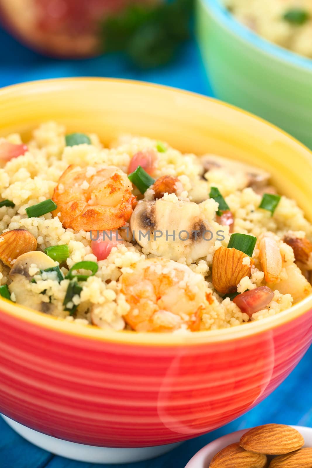 Couscous dish with shrimps, mushroom, almond, pomegranate seeds and green onion served in colorful bowl (Selective Focus, Focus on the tail of the shrimp on the top of the meal and on the mushroom slice next to it) 