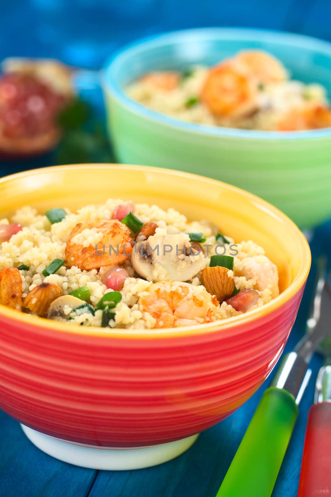 Two colorful bowls of couscous dish with shrimps, mushroom, almond, pomegranate seeds and green onion with forks on the side (Selective Focus, Focus on the tail of the shrimp on the top of the meal) 