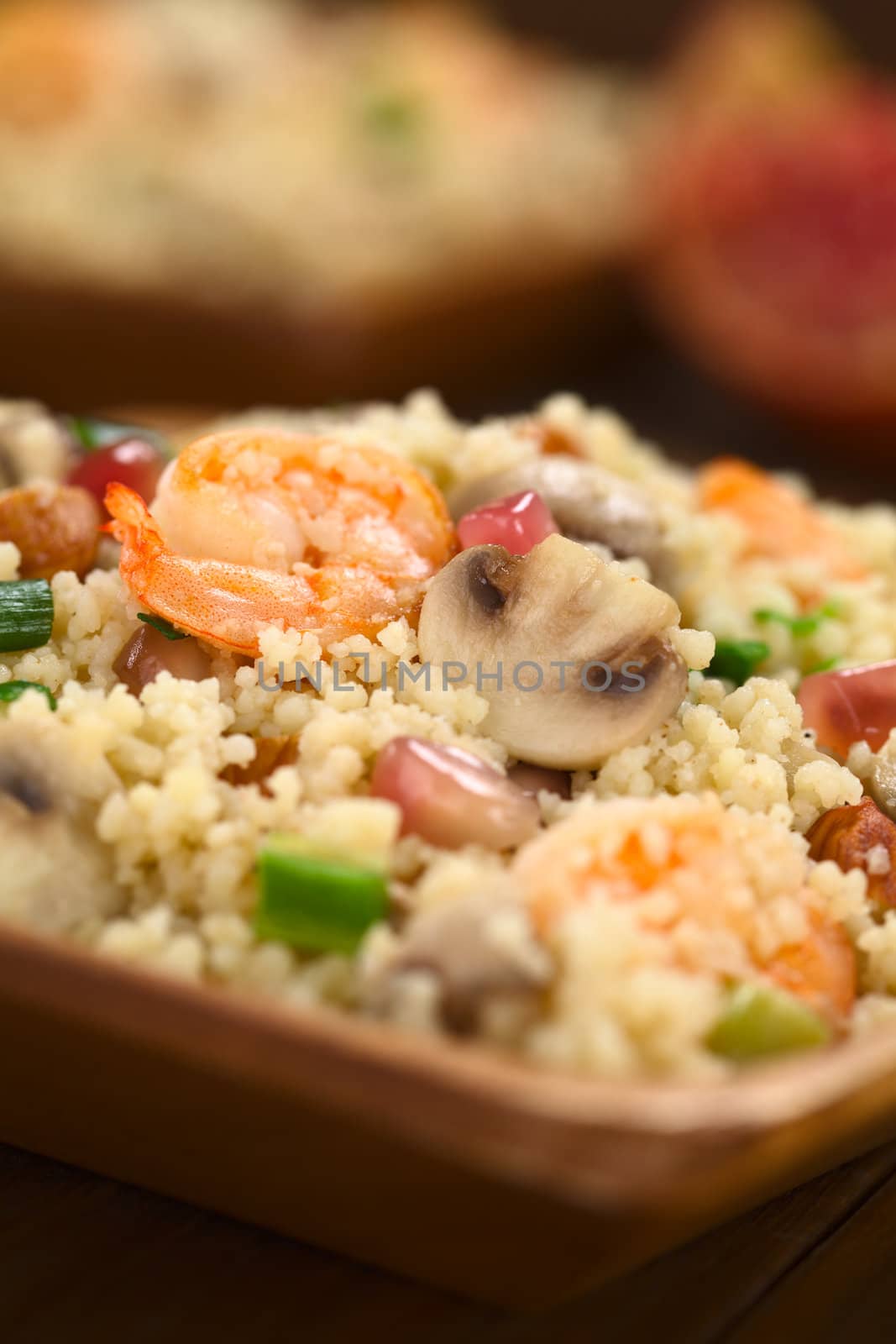 Couscous dish with shrimps, mushroom, almond, pomegranate seeds and green onion served on wooden plate (Selective Focus, Focus on the tail of the shrimp on the top of the meal and on the upper part of the mushroom slice next to it) 