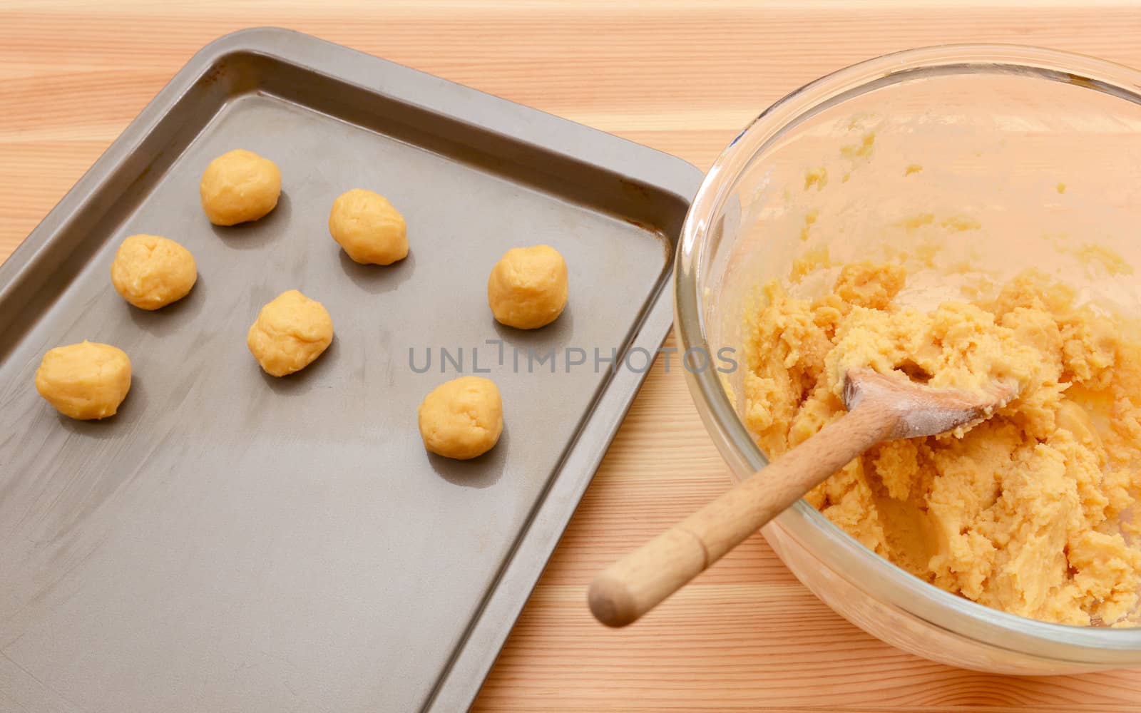 Balls of cookie dough on a baking sheet with bowl of mixture and wooden spoon