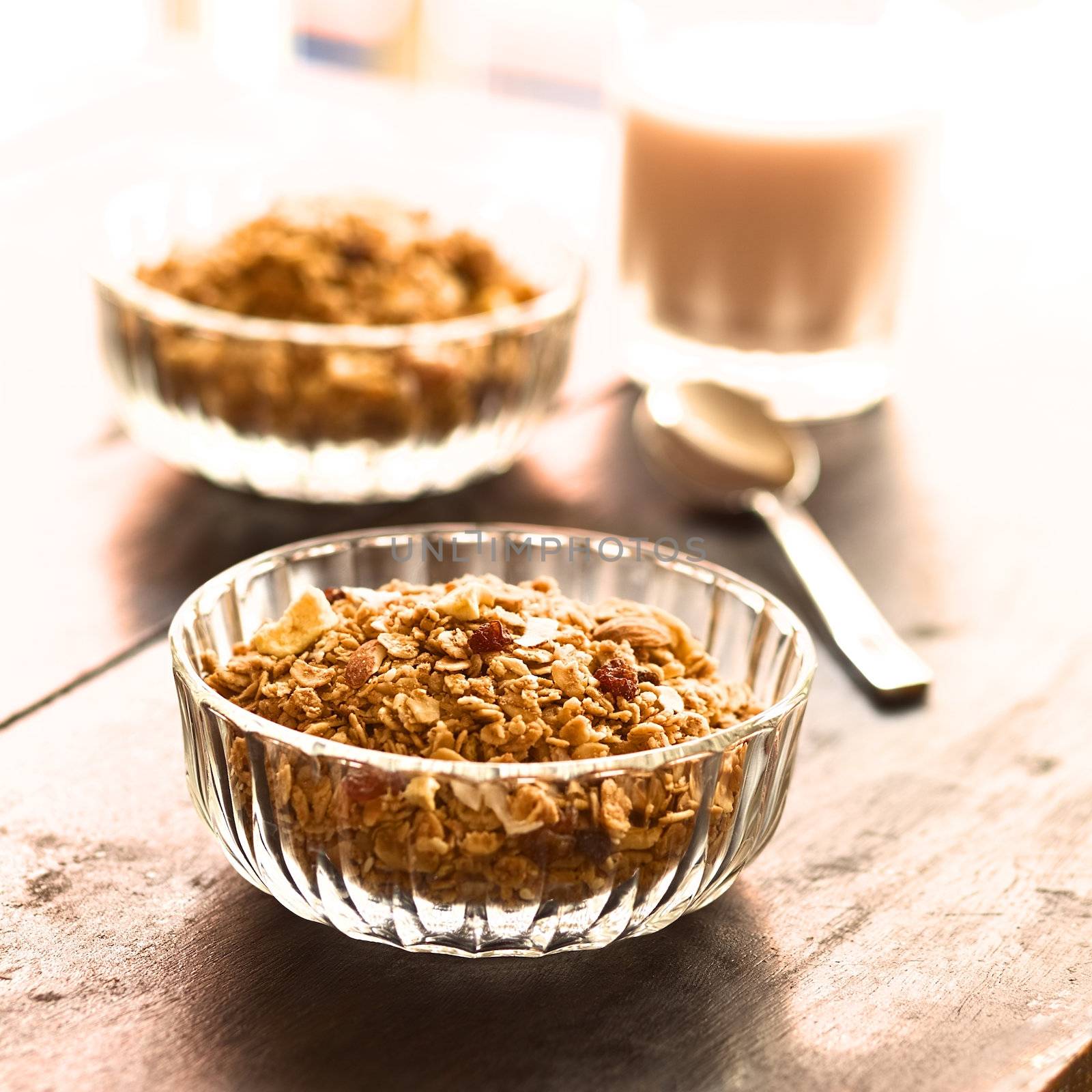 Breakfast cereal out of oat bran flakes, sesame, honey, almonds and dried fruits (coconut, apple, banana, raisins) in glass bowl with a glass of milk in the back (Selective Focus, Focus on the raisins on the top of the cereal)