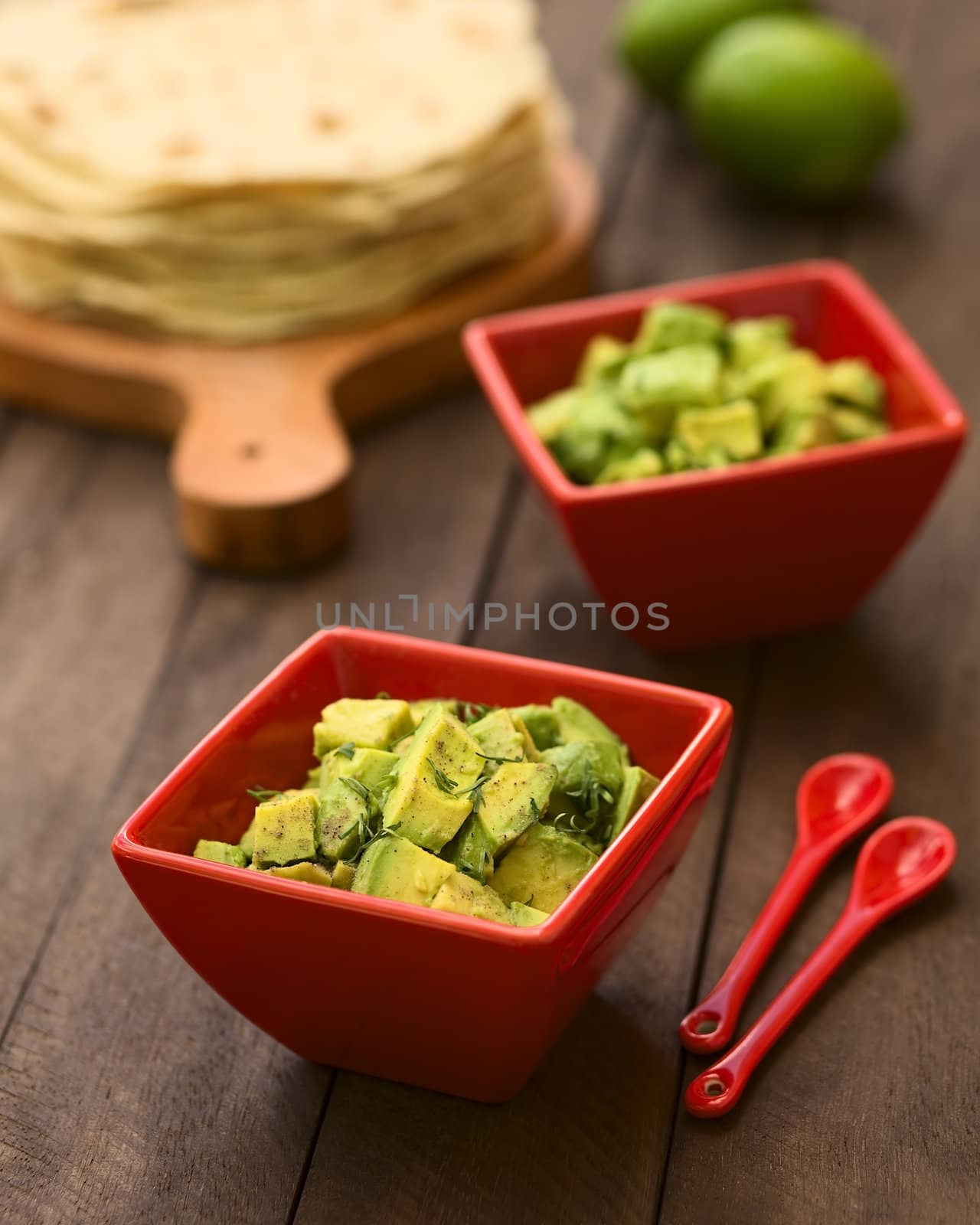 Two red bowls of fresh avocado salad prepared with lime juice, pepper, salt and garnished with fresh coriander leaves, homemade tortillas in the back (Selective Focus, Focus in the middle of the salad)  