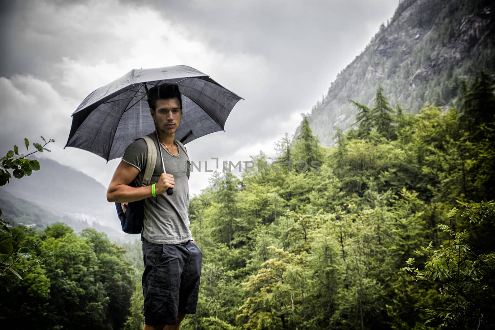 Smiling handsome young man holding an umbrella on a stormy day as he takes a walk along a trail in the mountains, looking at camera