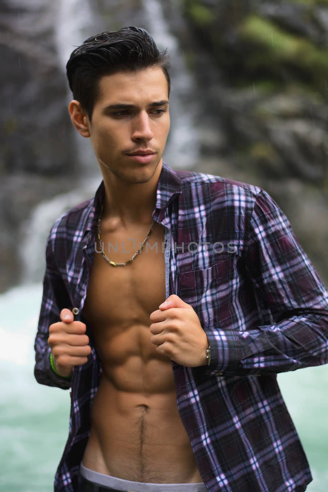 Handsome young man near mountain waterfall with open shirt, ripped athletic body