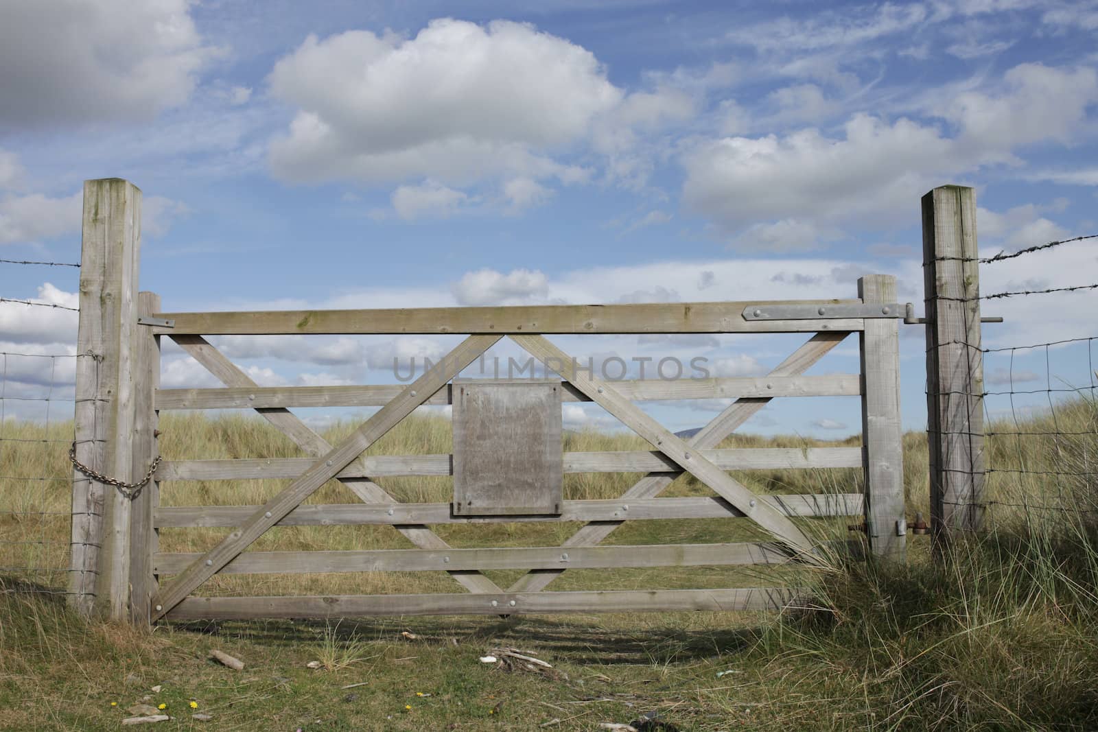 A closed wooden gate with a blank wooden sign against a grass landscape and blue sky with cloud.