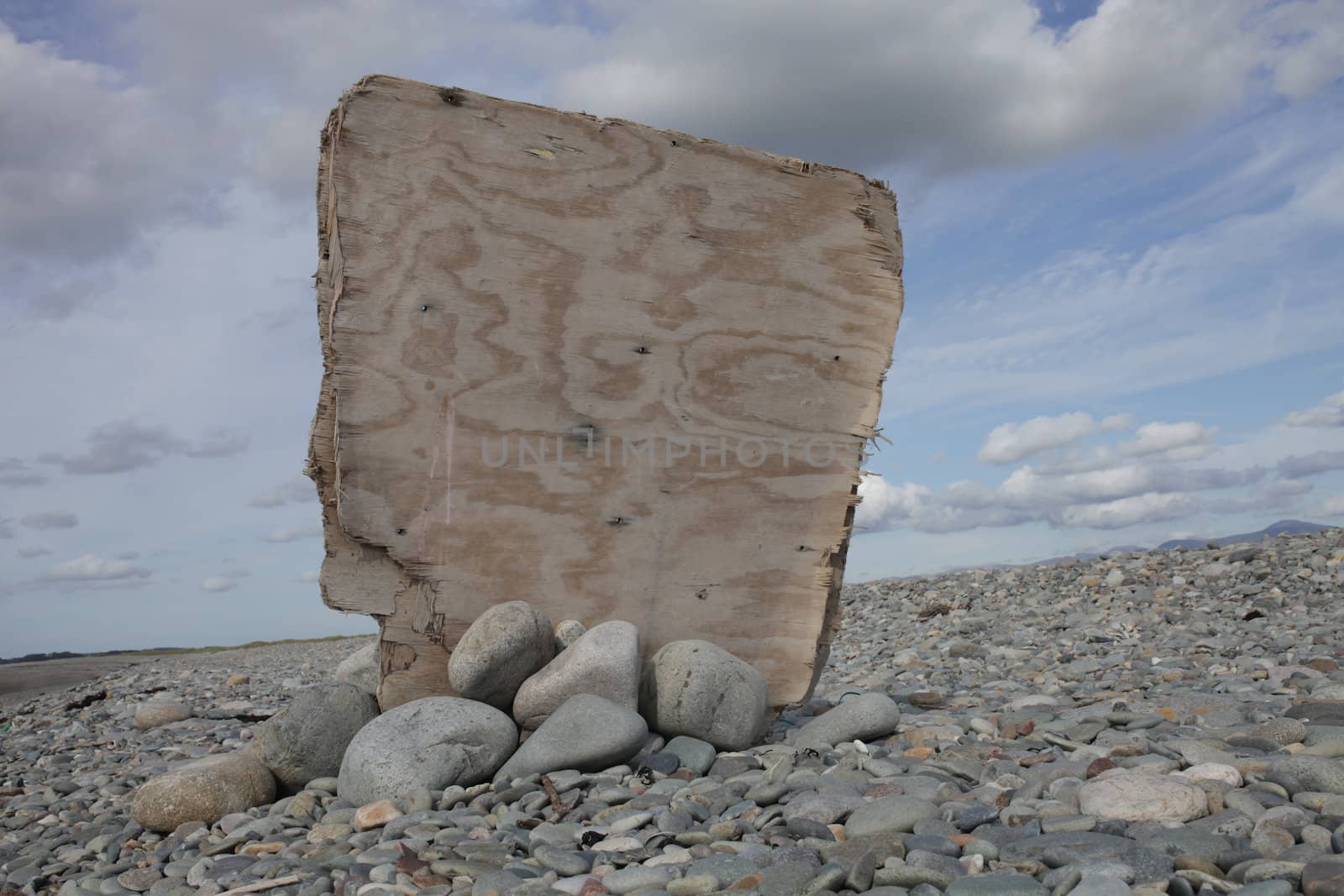 A blank plywood noticeboard standing upright propped by pebbles with a blue cloudy sky in the background.