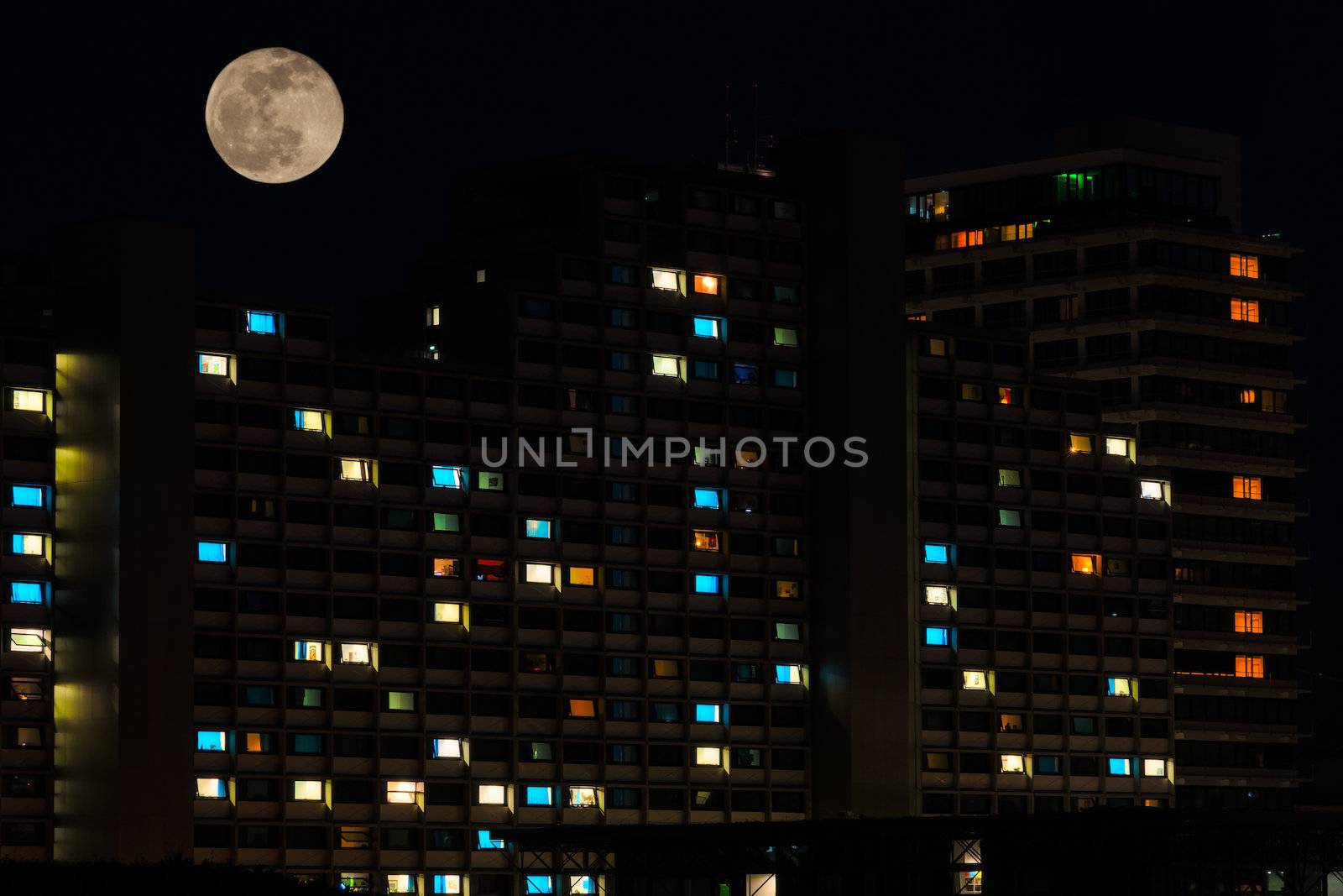 Full moon in night sky over colorful illuminated windows of residential high rise apartments house