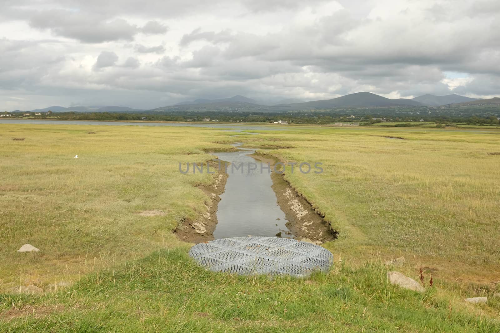 A circular metal drainage grill, hatch, on a grass mound leading to a stream crossing a tidal salt marsh.