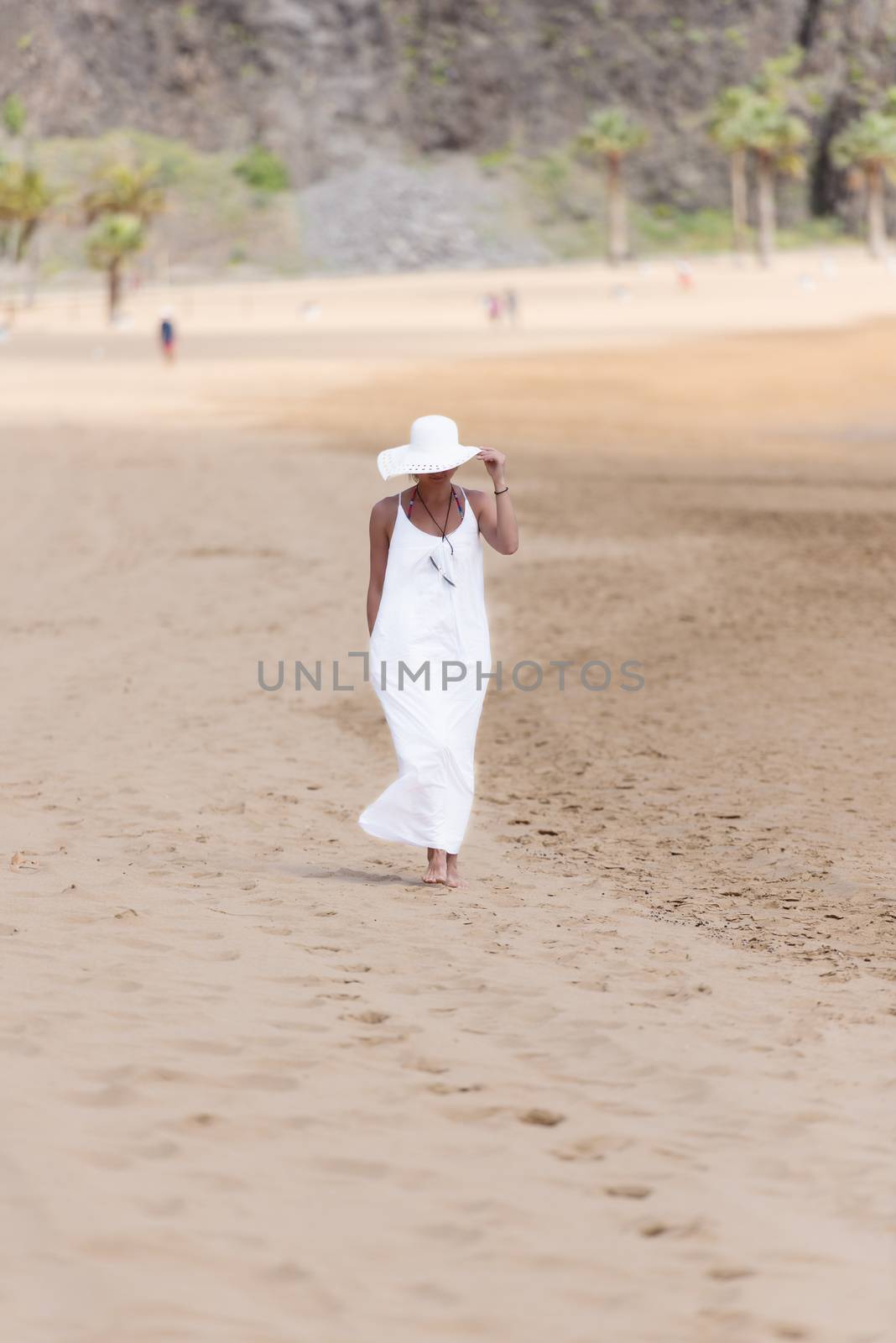 Barefoot girl in white hat and dress walking on a beach by Nanisimova