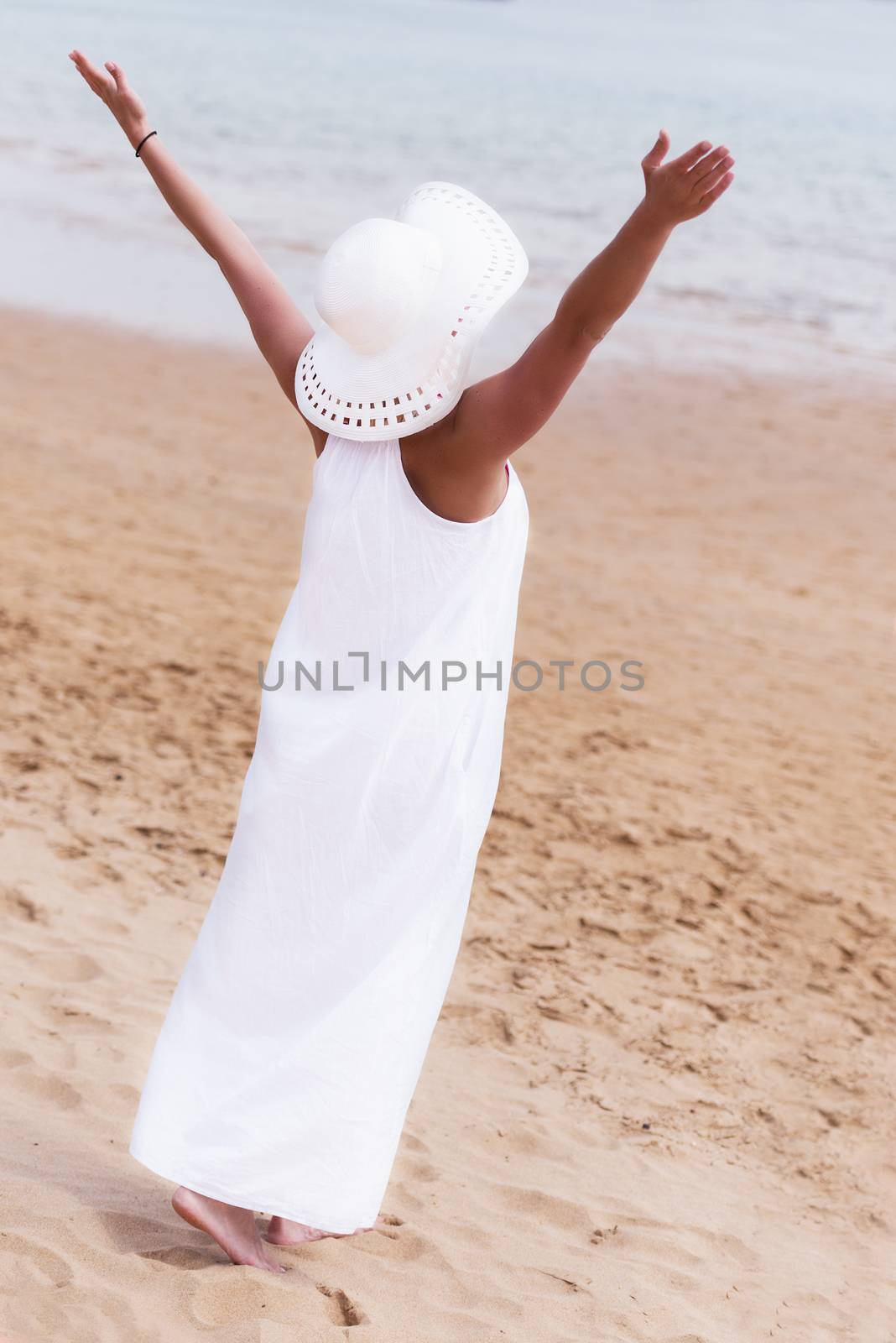 Lone girl in white dress and hat standing on beach with hands up