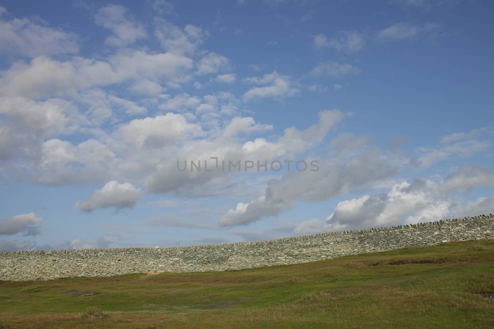 Green grass leads to an old dry stone wall built from white stone with a blue cloud sky in the distance.