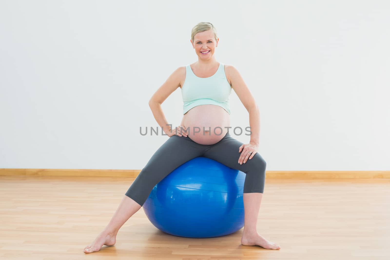 Smiling blonde pregnant woman sitting on blue exercise ball in a fitness studio