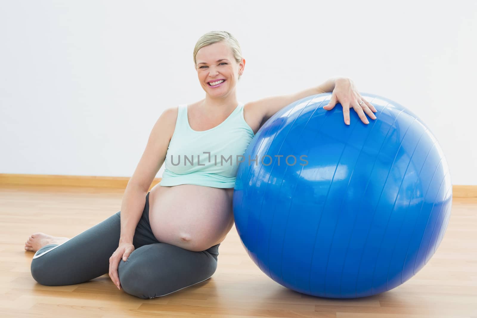 Pregnant woman sitting beside exercise ball smiling at camera in a fitness studio
