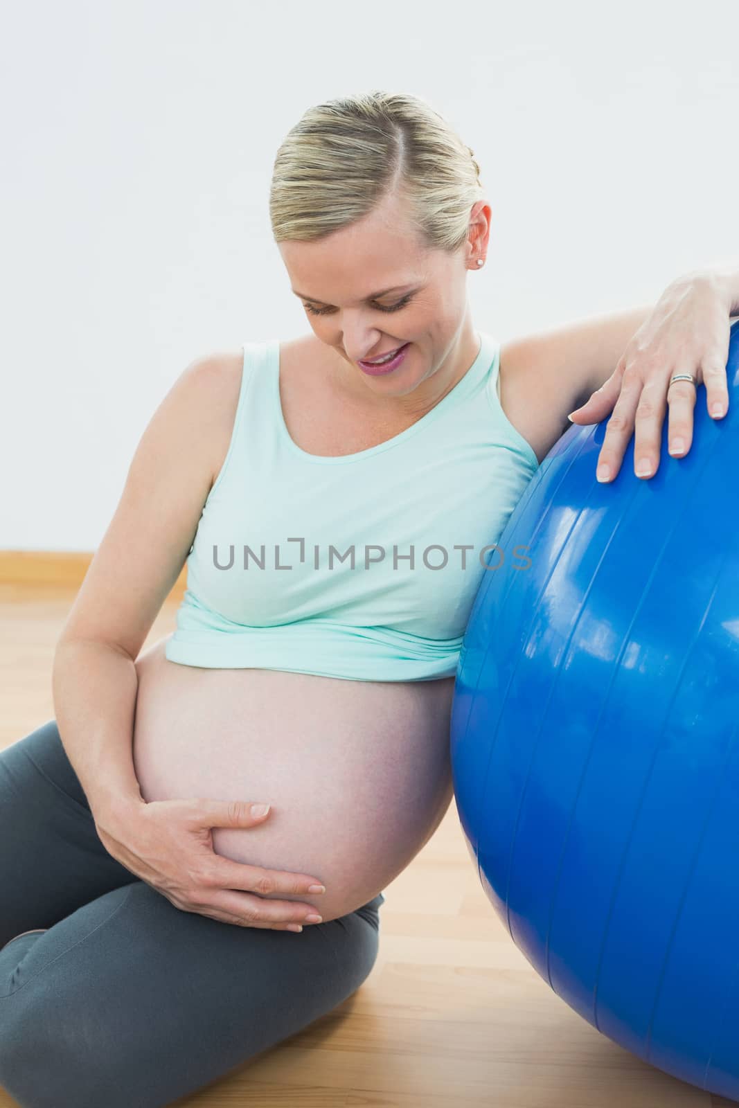 Smiling pregnant woman leaning against exercise ball holding her belly in a fitness studio