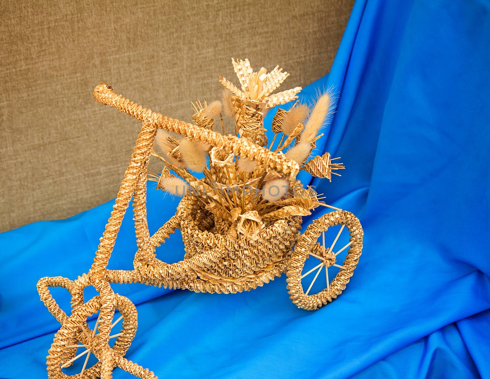 Souvenir product: the children's bicycle with a bouquet of flowers. It is made of wattled straw. It is presented against blue silk.