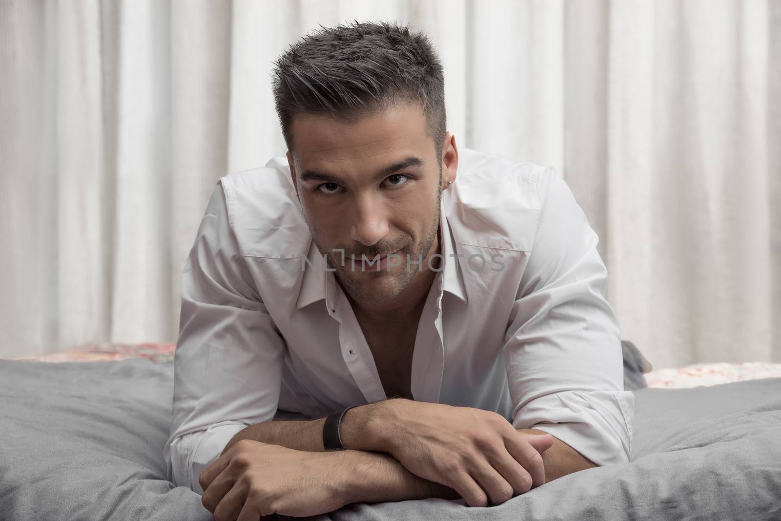 Sexy male model lying alone on his bed in his bedroom, looking at camera with a seductive attitude
