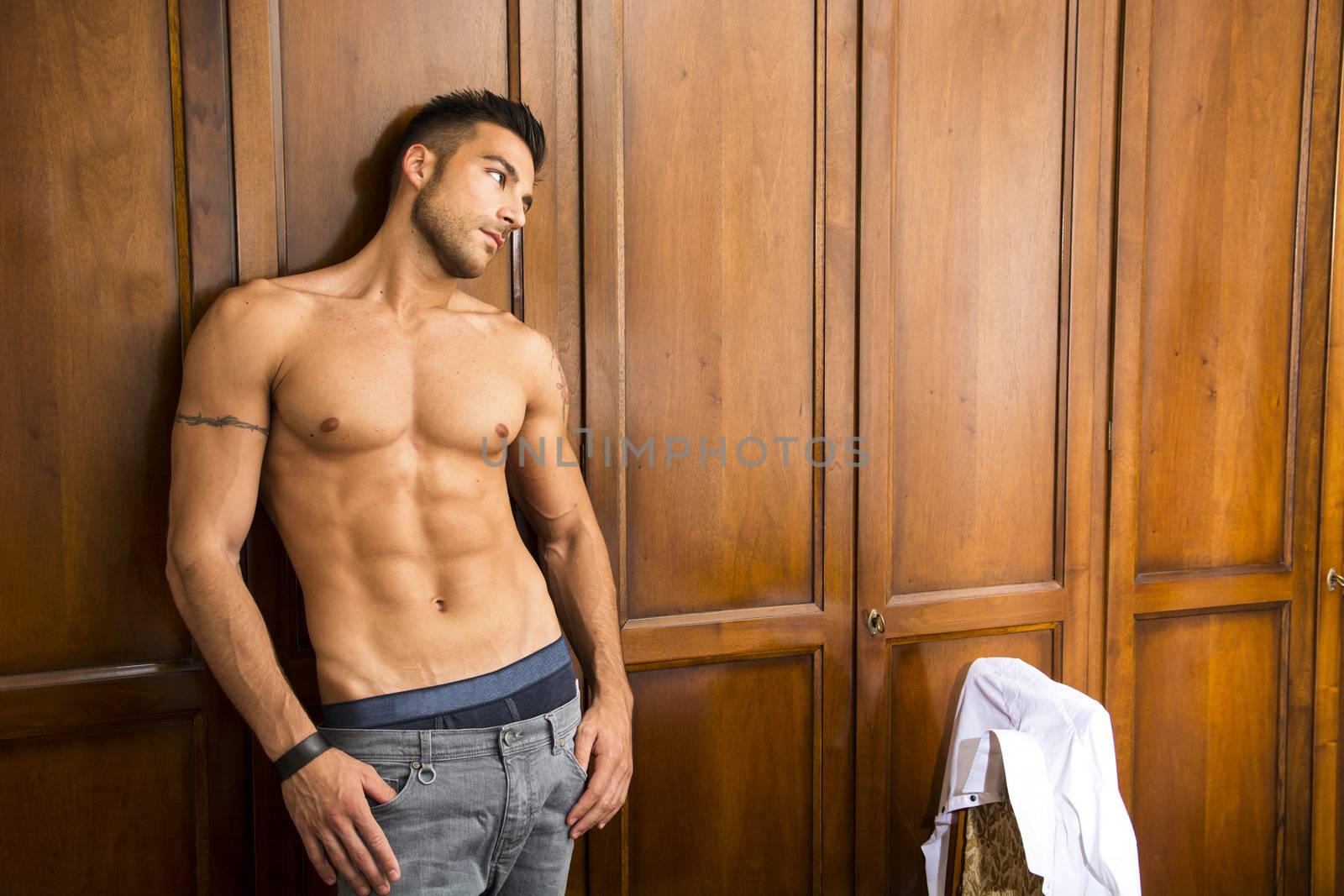 Sexy handsome young man standing shirtless in his bedroom with a shirt draped over chair with a smile