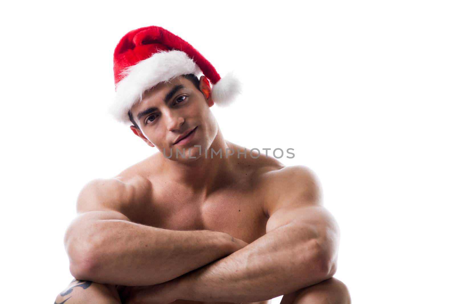 Attractive young muscle man sitting and smiling with muscular ripped body in Santa Claus's red hat