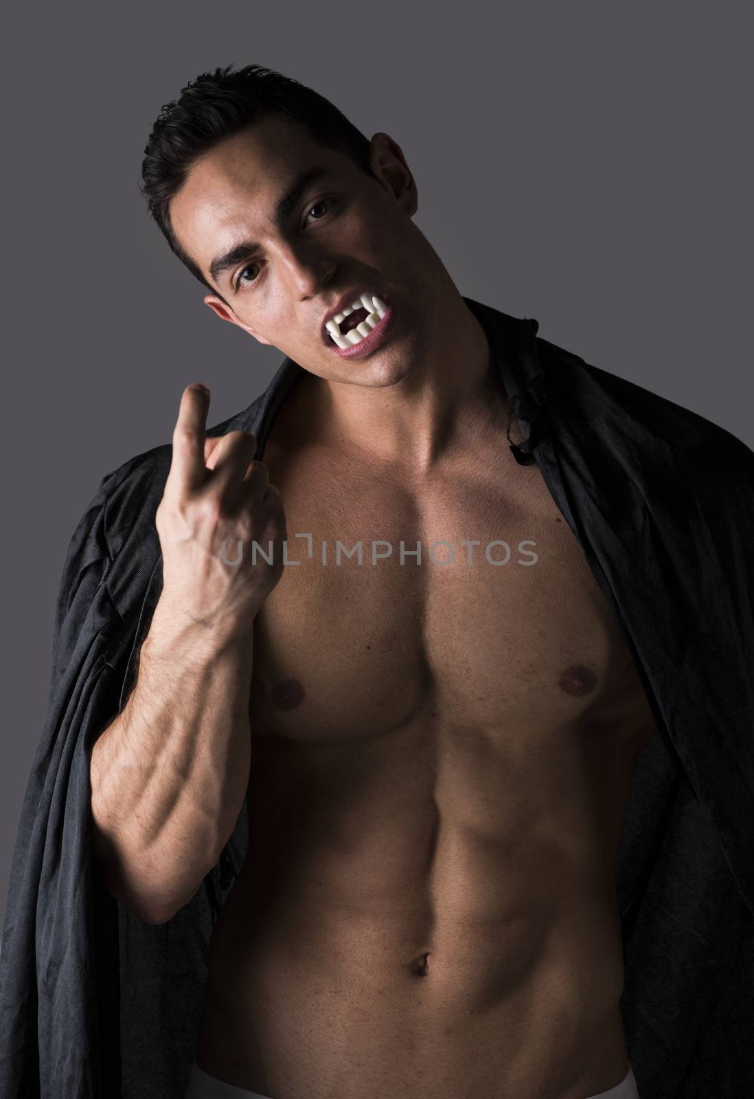 Naked muscular fit young man in briefs posing as a vampire or Dracula in a black cloak showing off his powerful body bearing his fangs with welcoming gesture