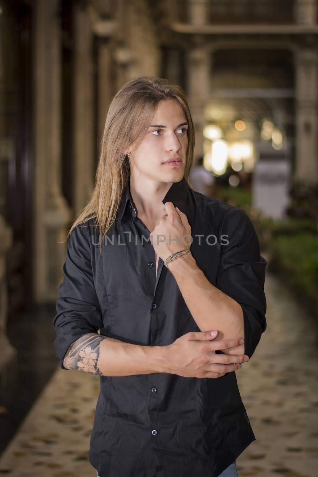 Handsome long hair man portrait in black shirt standing in elegant gallery in european city center