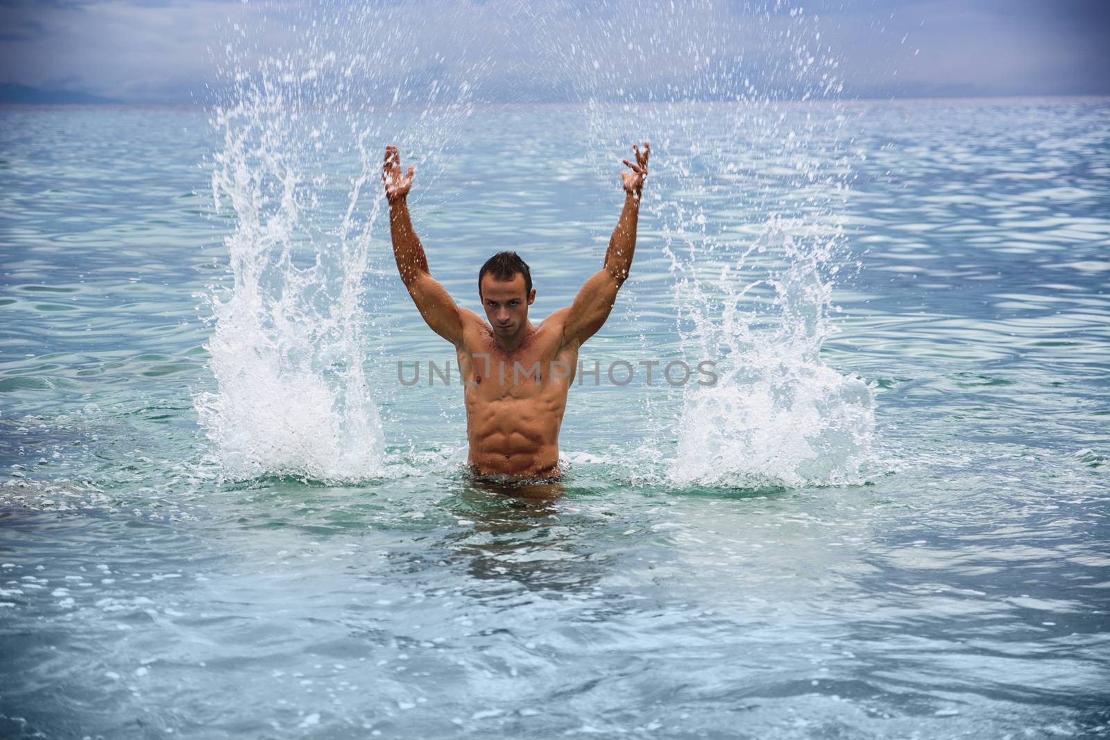 Handsome, hot young bodybuilder in the sea, splashing water up, showing his muscular torso and arms