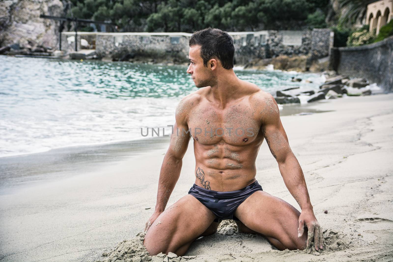 Attractive and fit young bodybuilder in bathing suit kneeling on the beach