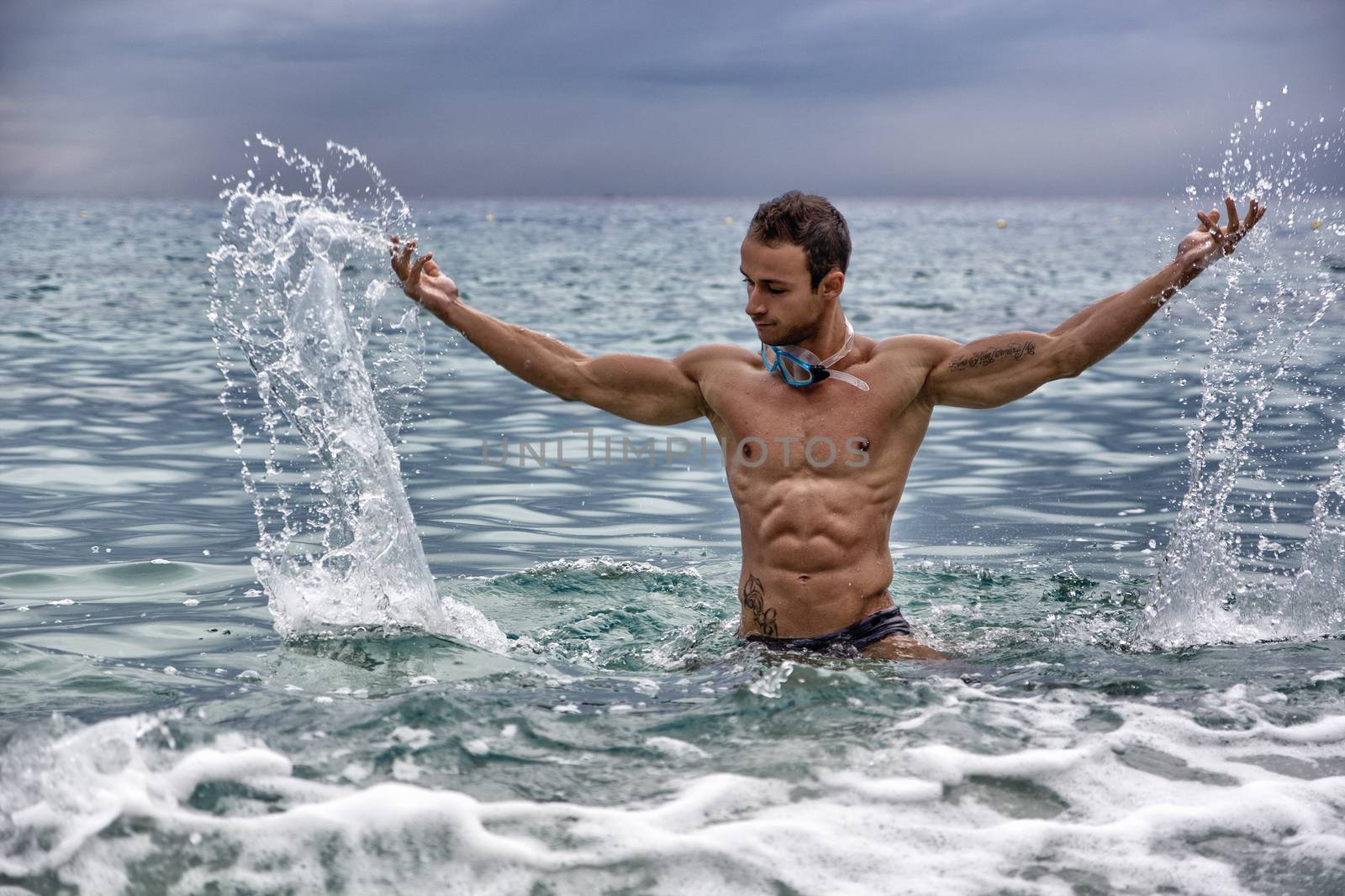 Handsome, hot young bodybuilder in the sea, splashing water up, showing his muscular torso and arms
