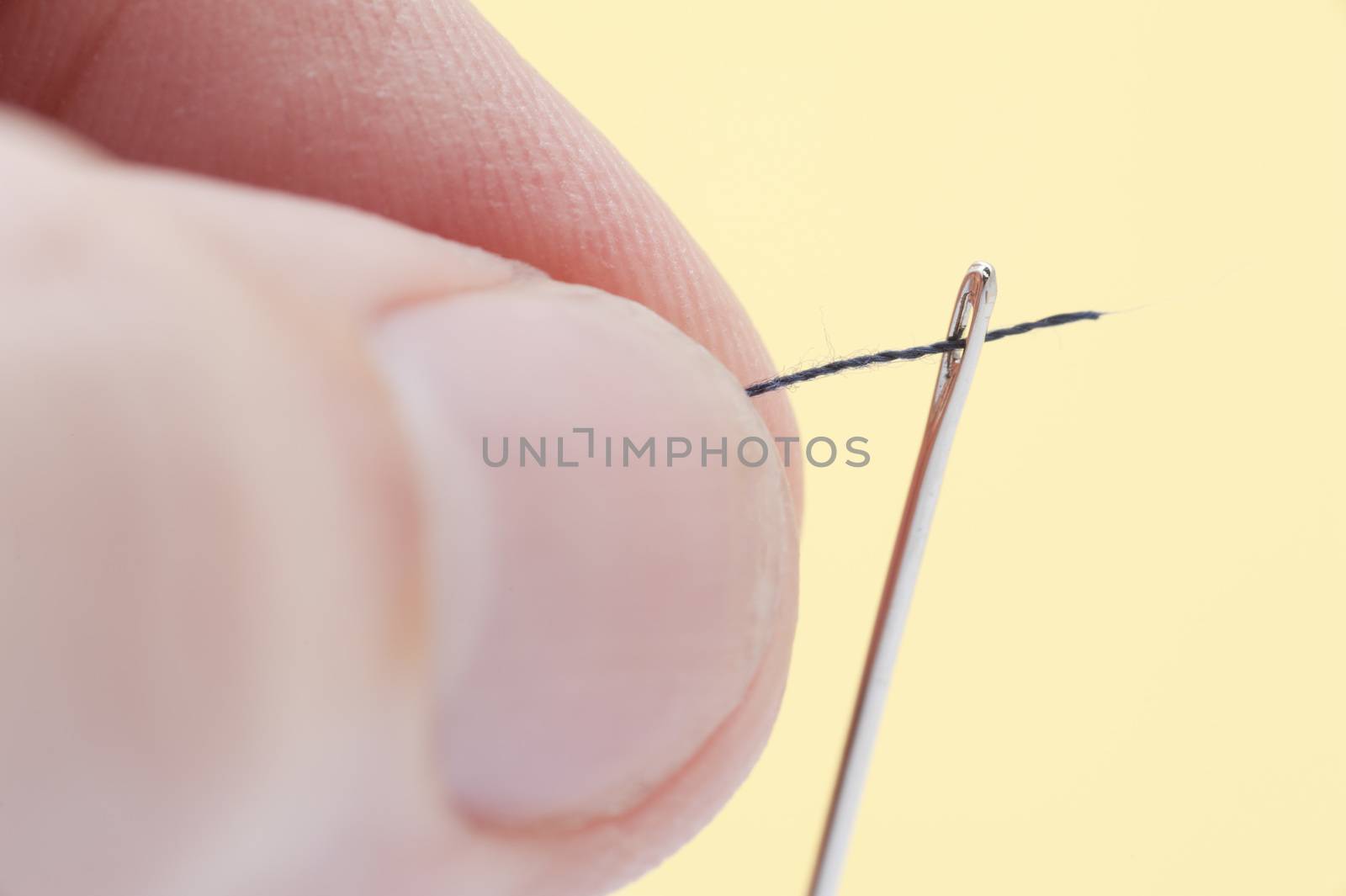 Close up view of a person threading a needle with yarn passing it through the eye over a yellow background in a needlework and handicraft concept