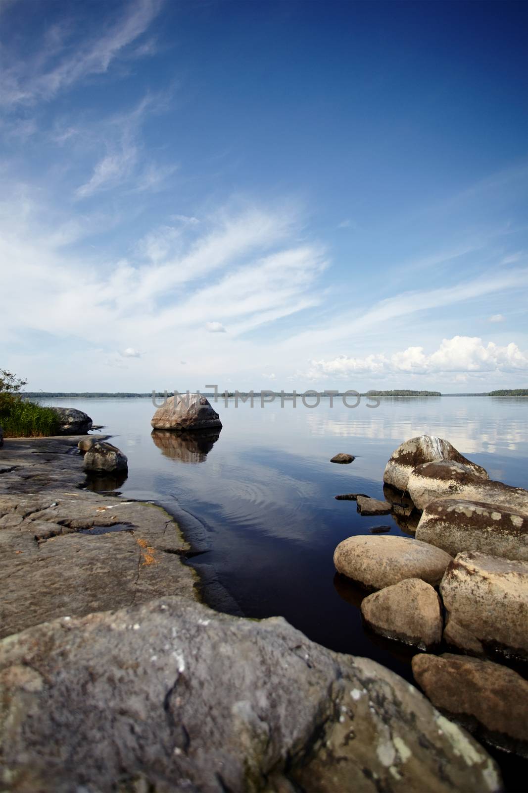 Water landscape with stones. Stones in water. The lake with stones. Beautiful landscape. Water smooth surface and the blue sky with clouds. Stone coast of the lake.