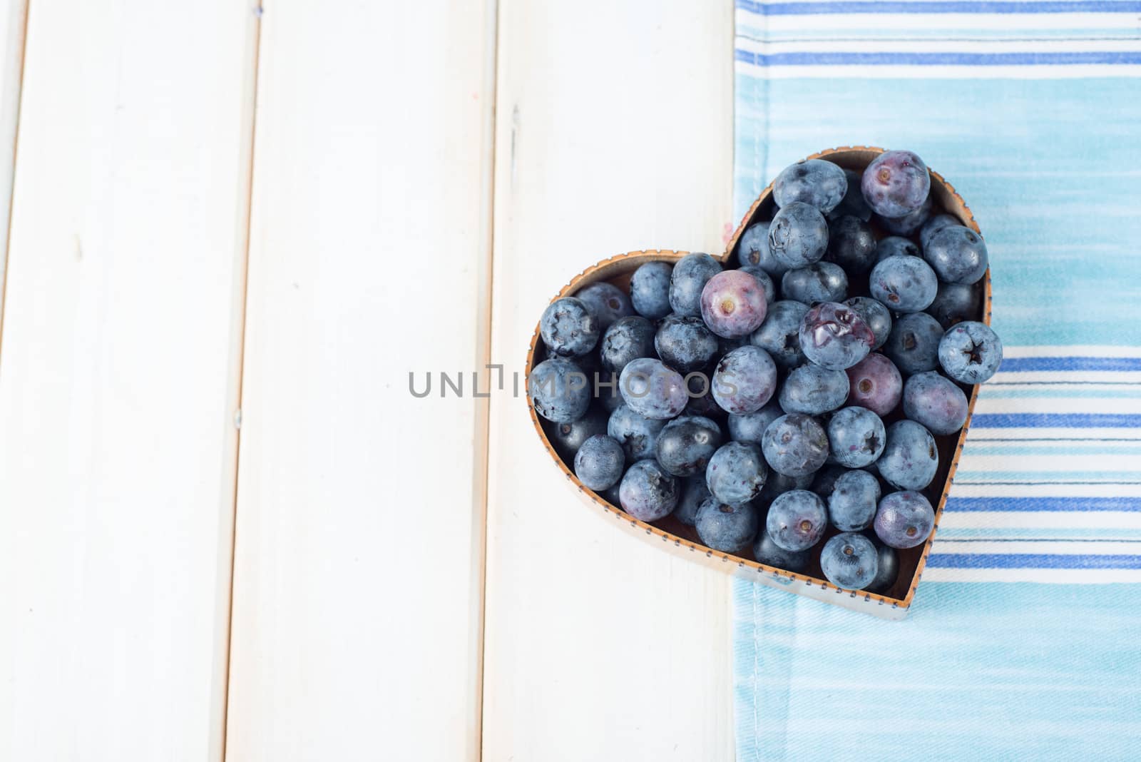 Fresh organic blueberries in heart style shape basket and blue kitchen cloth on white background retro kitchen table