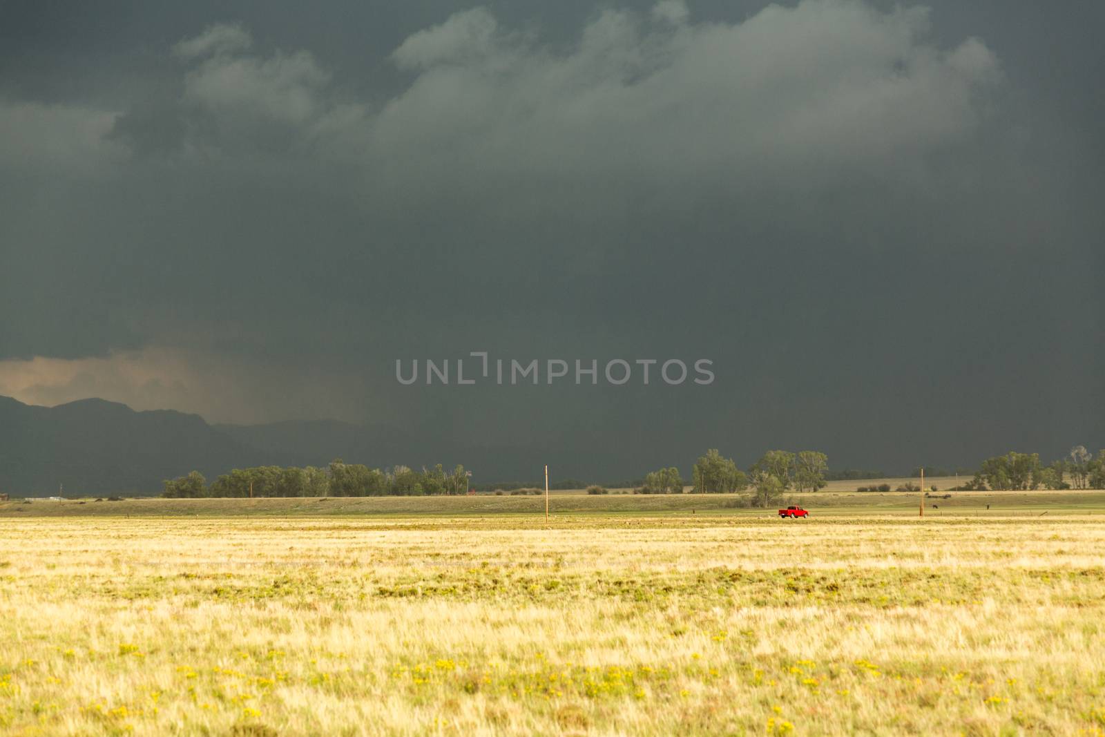 Sun illuminating prairie grassland with dark and forbidding thunderclouds and downpour with red van riding along lonely country road