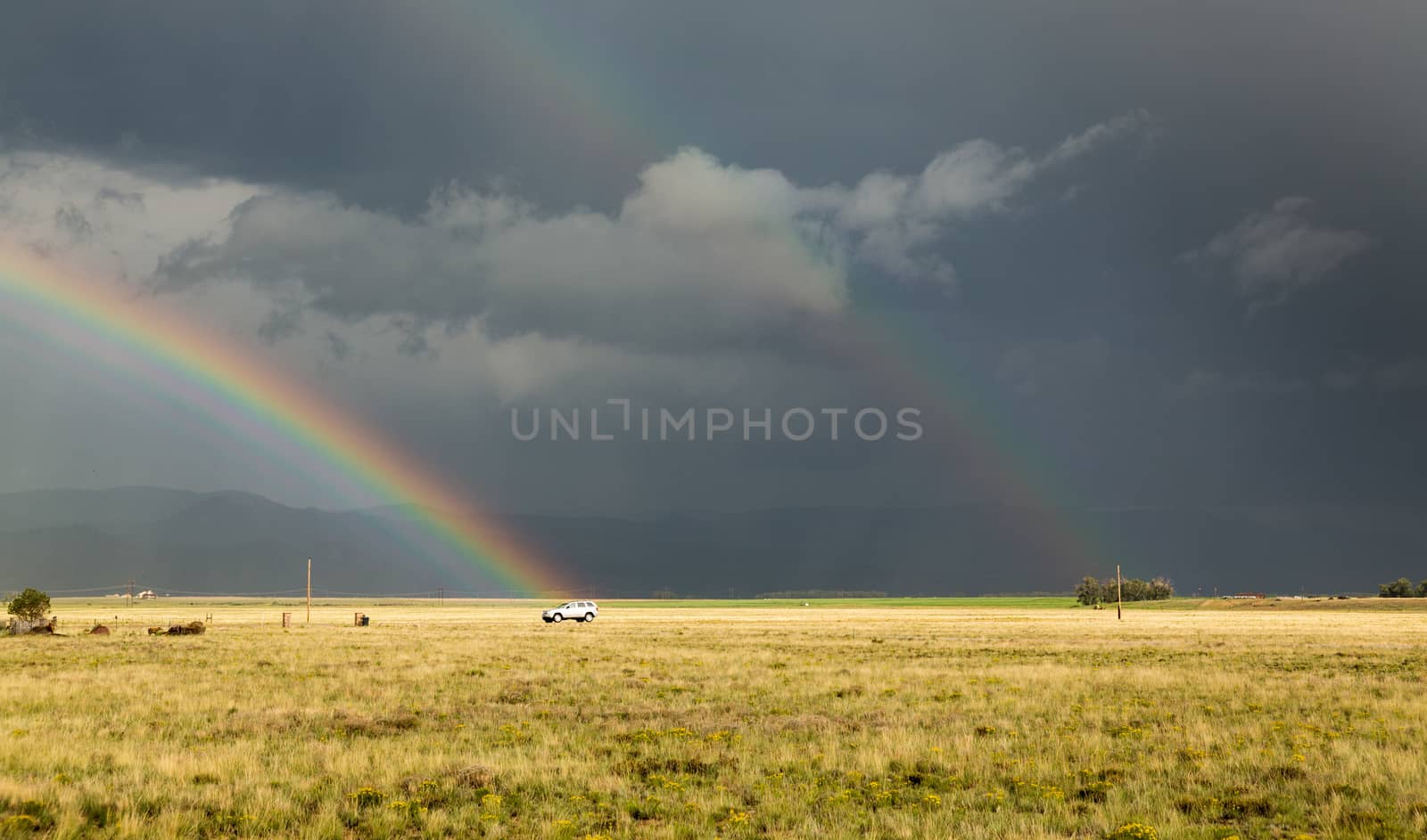 Sun illuminating prairie grassland with dark and forbidding thunderclouds and downpour with van riding along lonely country road and rainbow ending over the vehicle