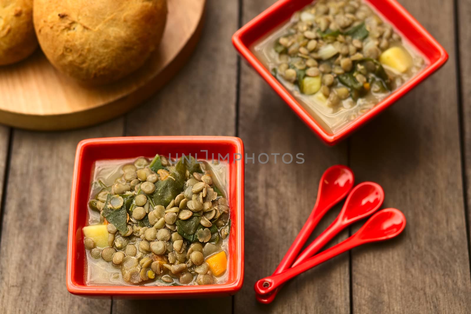 Vegetarian soup made of lentils, spinach, potato, carrot and onion served in red bowls with buns on the side (Selective Focus, Focus in the middle of the first soup)