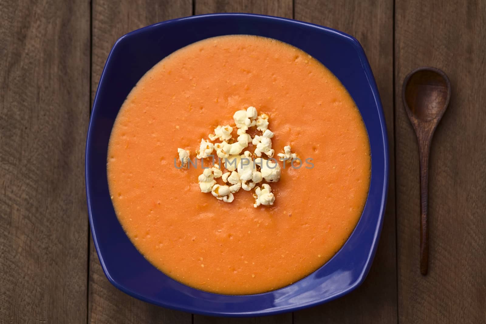 Ecuadorian tomato and potato cream soup served with popcorn on top in a blue soup plate with wooden spoon on the side (Selective Focus, Focus on the popcorn and on the lower part of the soup)