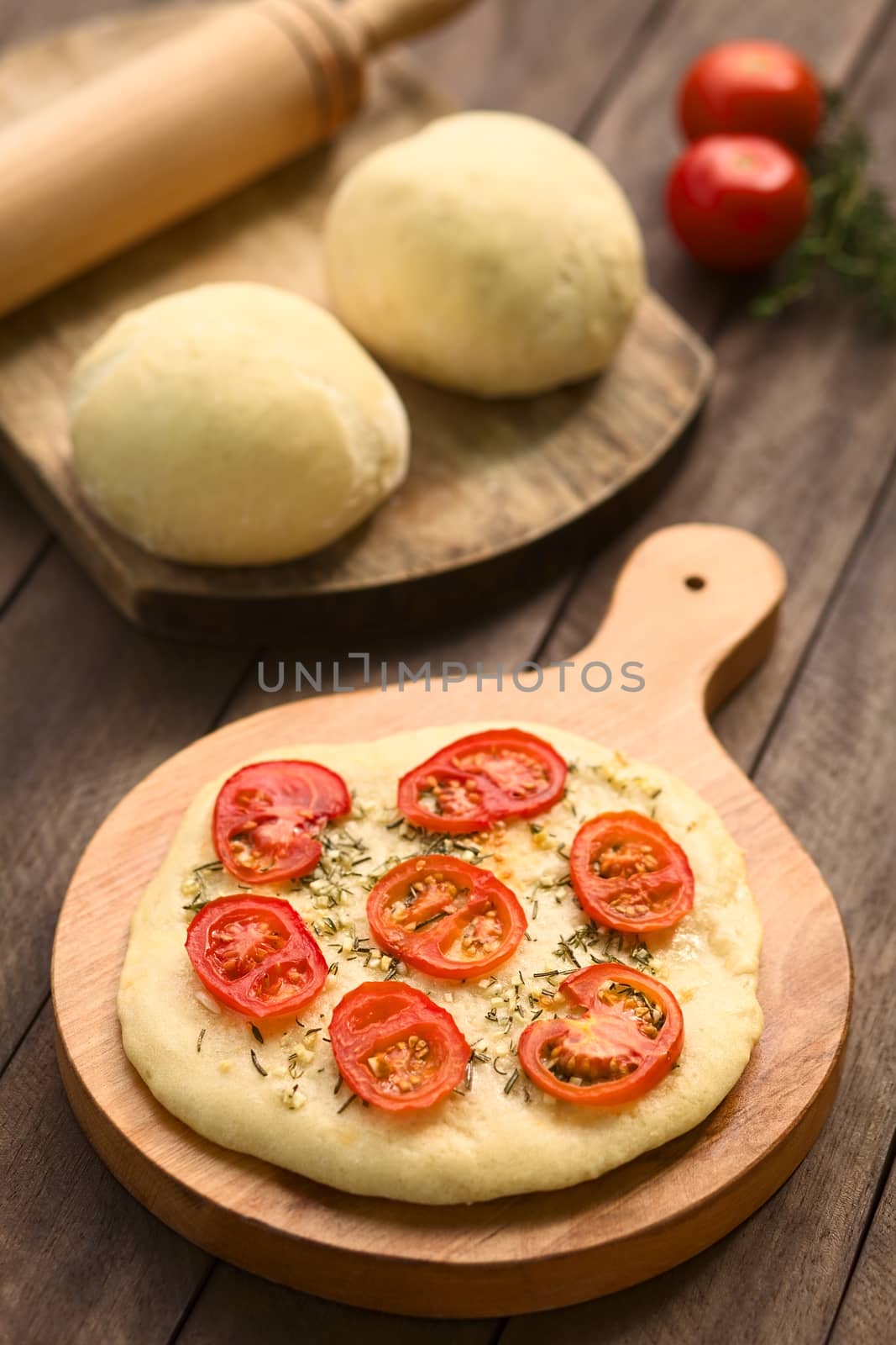 Freshly baked homemade flatbread made of a yeast dough topped with tomato slices, garlic and thyme leaves served on wooden board with balls of yeast dough in the back (Selective Focus, Focus on the front of the tomato slice in the middle)  