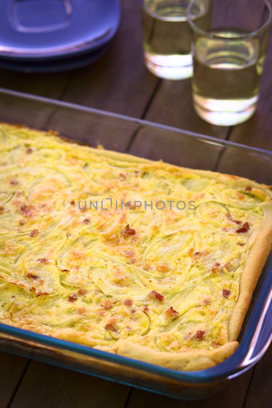 Traditional German Zwiebelkuchen or onion cake, made of a yeast dough and topped with onions and bacon in cream sauce in a glass baking pan, with white wine in the back (Very Shallow Depth of Field, Focus one third into the image) 
