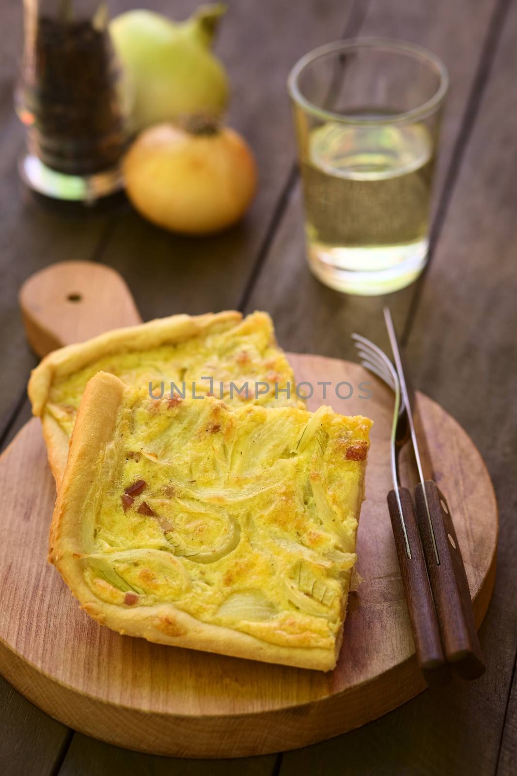 Pieces of traditional German Zwiebelkuchen or onion cake, made of a yeast dough and topped with onions and bacon in cream sauce with cutlery on wooden board, white wine in the back (Selective Focus, Focus one third into the image) 