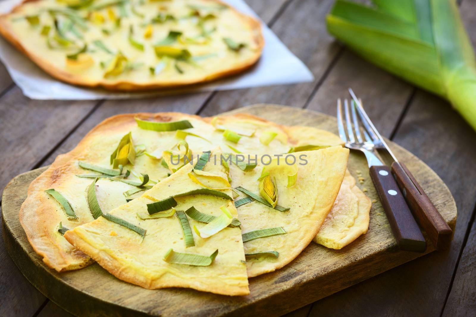 French vegetarian Tarte Flambee made of thin bread dough with a cream and leek topping, served on wooden board with knife and fork (Selective Focus, Focus two thirds into the first piece)  