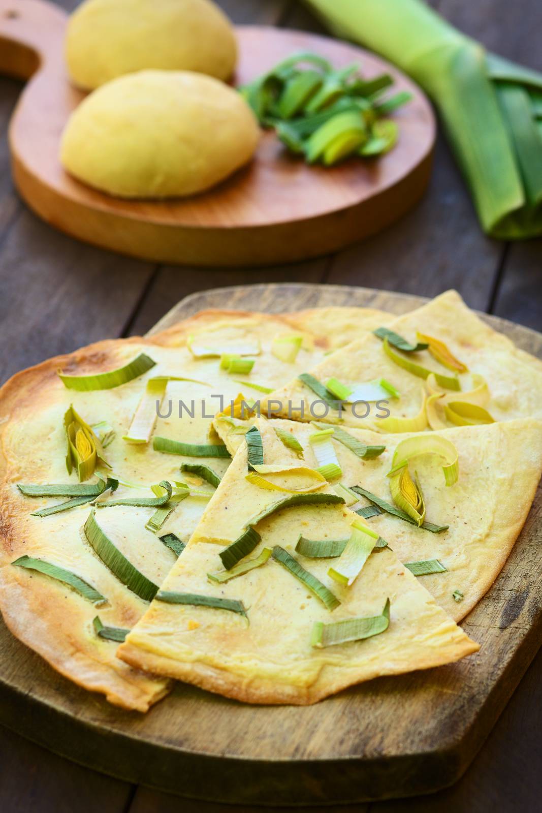 French vegetarian Tarte Flambee made of thin bread dough with a cream and leek topping, served on wooden board with ingredients in the back (Selective Focus, Focus on the upper part of the first piece)  