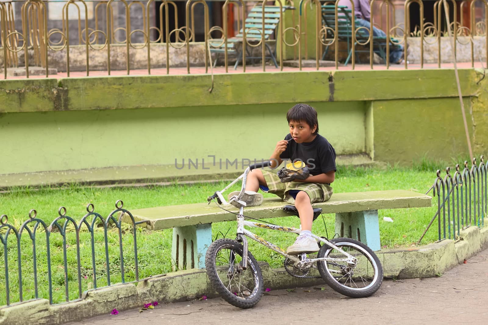 BANOS, ECUADOR - FEBRUARY 22, 2014: Unidentified boy with bike eating a snack while sitting on a bench in Sebastian Acosta Park on February 22, 2014 in Banos, Ecuador. 