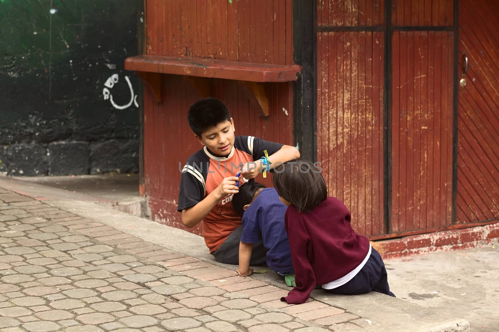 BANOS, ECUADOR - FEBRUARY 25, 2014: Unidentified boy cutting the hair of another child on the pavement on February 25, 2014 in Banos, Ecuador.  