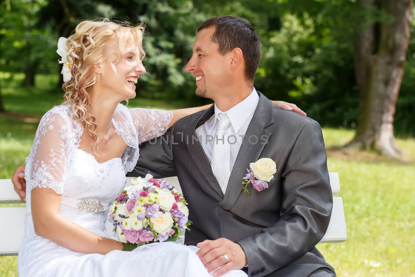 beautiful young wedding couple, blonde bride with flower and her groom looking to each other on park bench